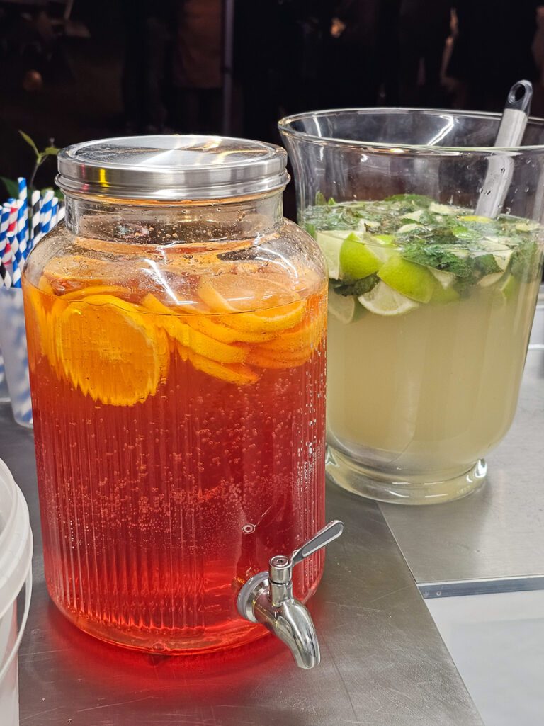 Un pichet contenant des tranches d'orange et une boisson rouge trône à côté d'un pichet de limonade agrémentée de citron vert et de menthe sur le comptoir, créant ainsi une présentation colorée, idéale pour tout camion-bar événementiel.