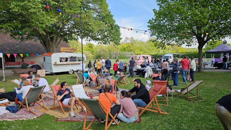 Les gens se détendent sur des transats et des couvertures de pique-nique lors d'un festival en plein air, profitant de l'ambiance animée d'une guinguette événementielle, avec des food trucks, des guirlandes lumineuses et des arbres en arrière-plan, par une journée ensoleillée.