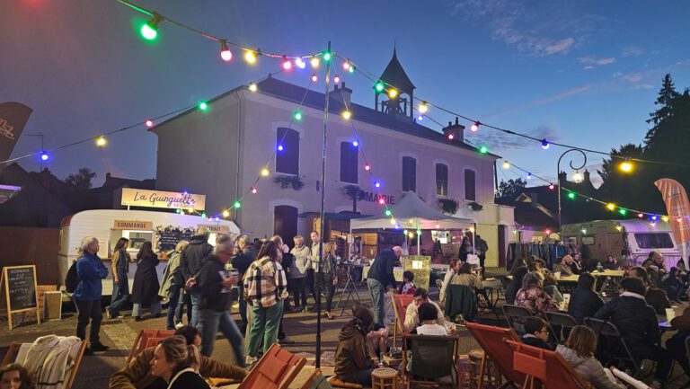 Un festival en plein air organisé en soirée, où les participants se rassemblent sous des guirlandes lumineuses colorées, au milieu des stands de restauration, avec un bâtiment blanc en arrière-plan. Dans cette guinguette événementielle animée, les invités sont assis, se promènent ou restent debout tout en profitant de l'ambiance animée.