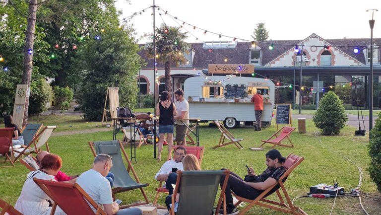 Des gens se détendent sur des transats posés sur l'herbe, près d'une guinguette événementielle — un food truck festif décoré de guirlandes lumineuses. Certains discutent, tandis que d'autres consultent leur téléphone. Des arbres et un bâtiment se dessinent en arrière-plan. L'ambiance est décontractée et conviviale.