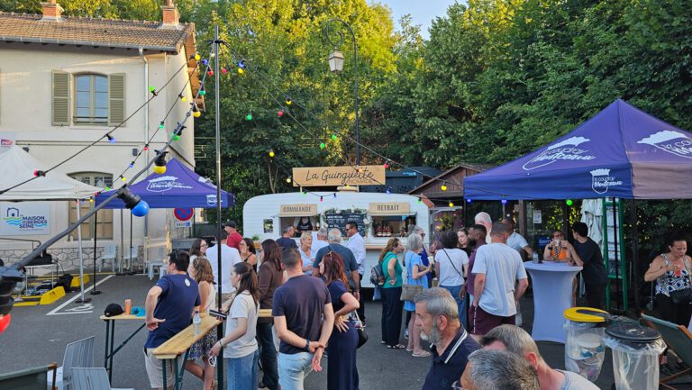 Une foule se rassemble devant un stand de restauration en plein air appelé « La Guinguette », entouré de lumières colorées, de tentes et d’arbres. Les gens discutent, commandent à manger et profitent de l’ambiance animée et festive de cette guinguette événementielle par une journée ensoleillée.
