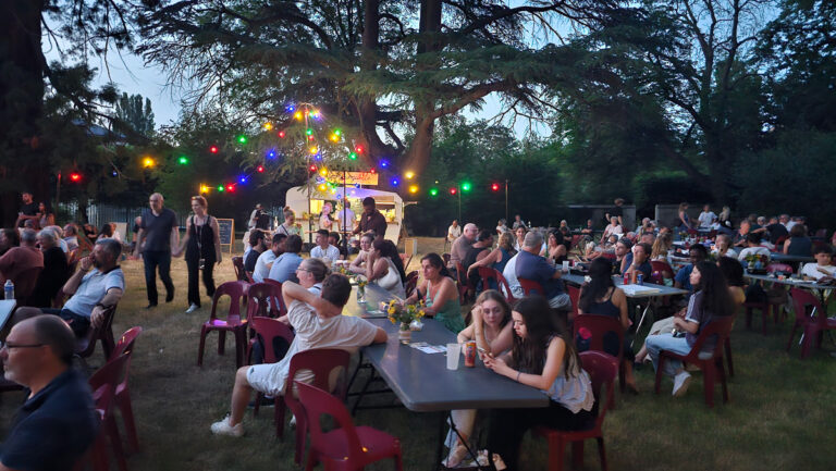 Le soir, les gens s'installent à des tables en terrasse, sous des guirlandes lumineuses colorées, pour déguster des plats et des boissons dans une guinguette événementielle animée. On aperçoit des arbres en arrière-plan et un food truck blanc.