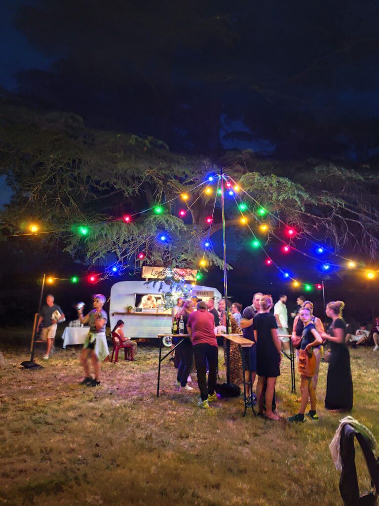 Le soir venu, les gens se rassemblent sous des guirlandes lumineuses colorées près d'un food truck, dans un espace vert en plein air, pour savourer un repas et discuter. L'ambiance est animée et festive, reflétant l'esprit joyeux d'une guinguette événementielle.