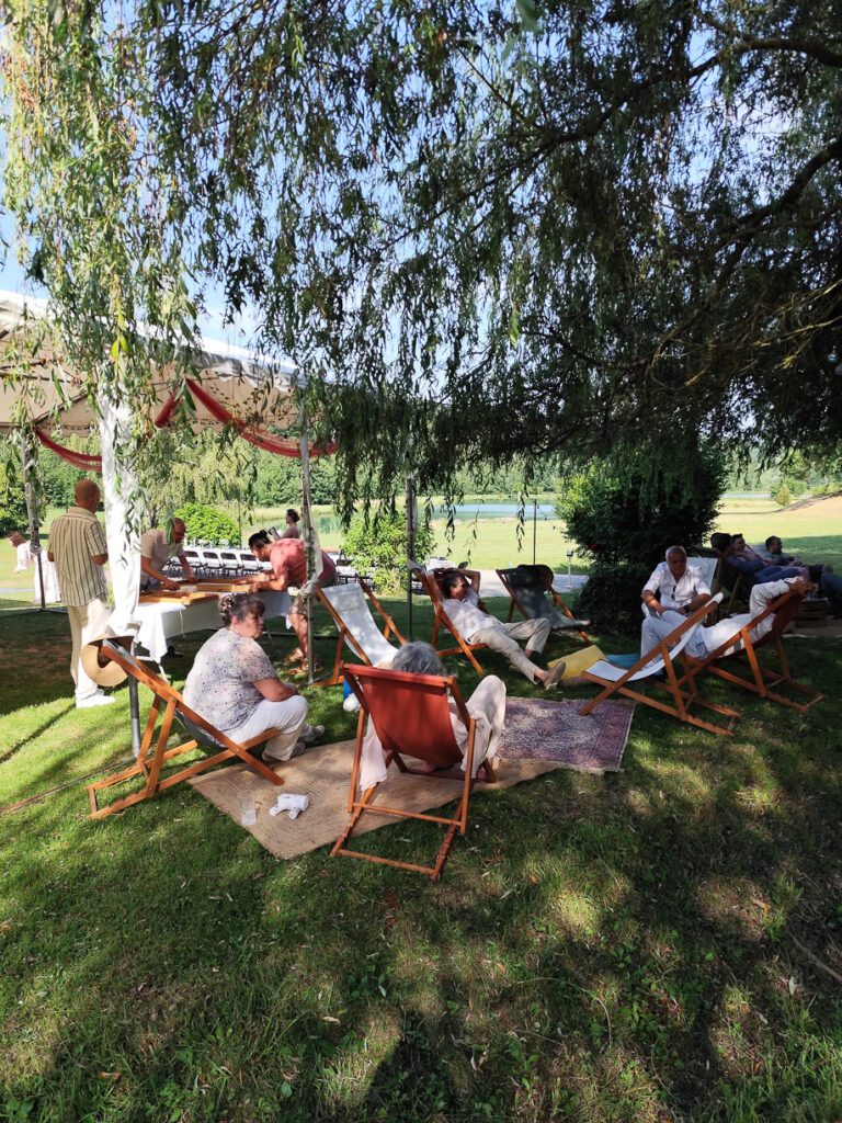 Les participants se détendent sur des transats à l'ombre d'un arbre lors d'un rassemblement en plein air, profitant de l'ambiance décontractée d'une guinguette événementielle. Certains sont assis sur des tapis, tandis que d'autres discutent à proximité. On aperçoit en arrière-plan un auvent, des tables et des prairies verdoyantes.