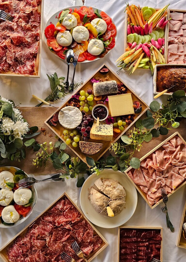 Un plateau de charcuterie haut en couleur composé d'un assortiment de viandes, de fromages, de légumes frais, de fruits, d'une miche de pain ronde et de salades caprese, disposé sur une nappe blanche et agrémenté de feuilles vertes et de fleurs, évoquant le charme festif d'une guinguette événementielle.