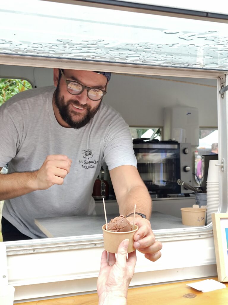 En Île-de-France, un homme souriant, portant des lunettes et un t-shirt gris, tend à un client une coupe de glace au chocolat accompagnée de deux bâtonnets à travers la vitrine d’un food truck événementiel.