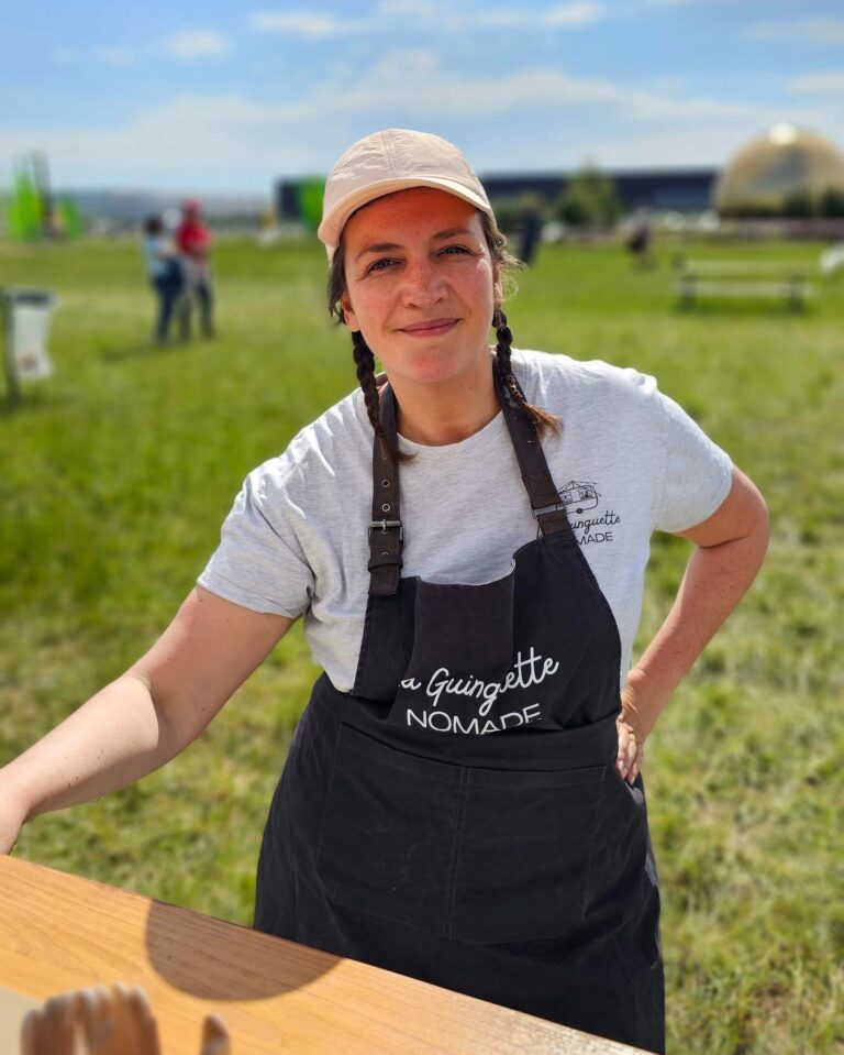 Une femme coiffée d'un chapeau blanc, vêtue d'un t-shirt gris et d'un tablier noir, se tient dehors sur l'herbe et sourit à la caméra. Derrière elle, des tables, des bancs et une structure en forme de dôme laissent entrevoir un événement animé autour de food trucks en Île-de-France, sous un ciel bleu.