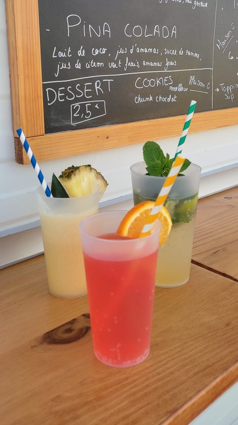 Trois boissons colorées servies dans des gobelets en plastique avec des pailles rayées trônent sur un comptoir en bois, l'idéal pour tout bar mobile événementiel. Elles sont agrémentées d'ananas, de menthe et d'une rondelle d'orange, tandis qu'un menu écrit à la craie est visible en arrière-plan.