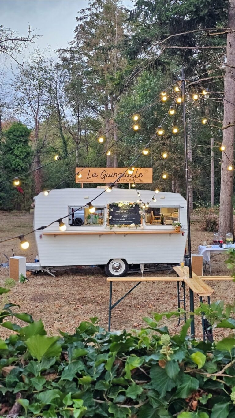 Un food truck blanc portant l'enseigne « La Guinguette » est garé sur la pelouse, au milieu des arbres, et décoré de guirlandes lumineuses. Dans l'esprit d'une guinguette événementielle, un comptoir en bois et quelques tables ont été installés à l'extérieur, invitant les convives à profiter de l'ambiance festive en plein air.