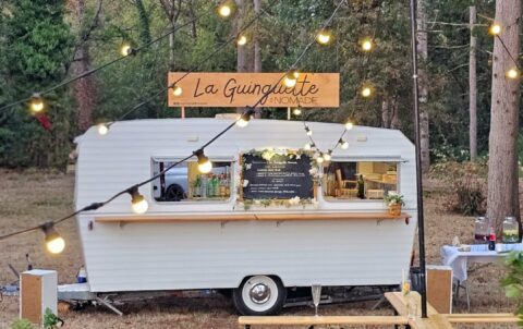 Un food truck blanc baptisé « La Guinguette » est garé en plein air, au milieu des arbres. Des guirlandes lumineuses sont suspendues au-dessus, créant une ambiance chaleureuse, idéale pour une guinguette événementielle. Le menu est affiché sur la vitrine du food truck, et des tables en terrasse sont disposées à proximité.