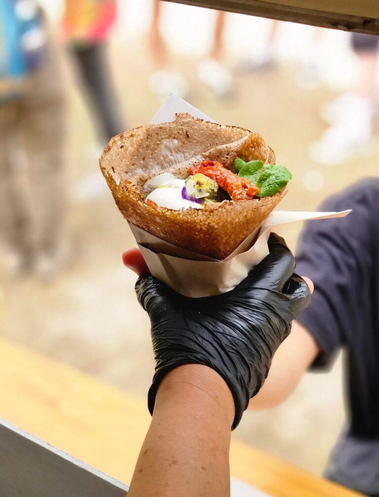 Une personne portant un gant noir tend à quelqu’un, au comptoir d’un food truck événementiel, une crêpe au sarrasin garnie de crème, de tomates rôties et de légumes verts.