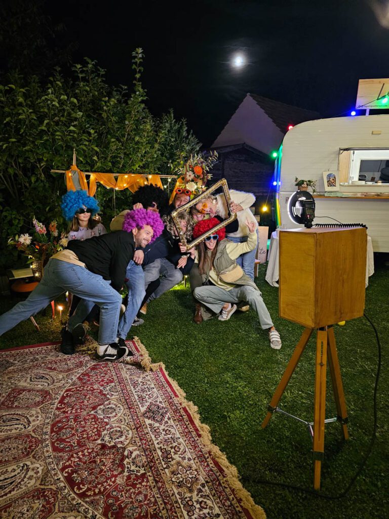 Quatre personnes, coiffées de perruques colorées et portant des lunettes de soleil, prennent la pose de manière enjouée avec un cadre photo, de nuit, à l'extérieur, près d'un food truck événementiel décoré et d'un appareil photo. Une lune éclatante et des guirlandes lumineuses illuminent cette scène festive.