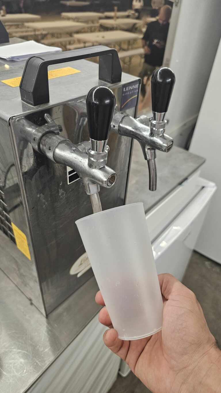 Une main tient un gobelet en plastique translucide sous un distributeur de boissons métallique muni de deux robinets, prête à remplir le gobelet. Le décor semble être celui d’une cafétéria ou d’un camion-bar événementiel, ce qui apporte une touche dynamique à l’ambiance du service de boissons.