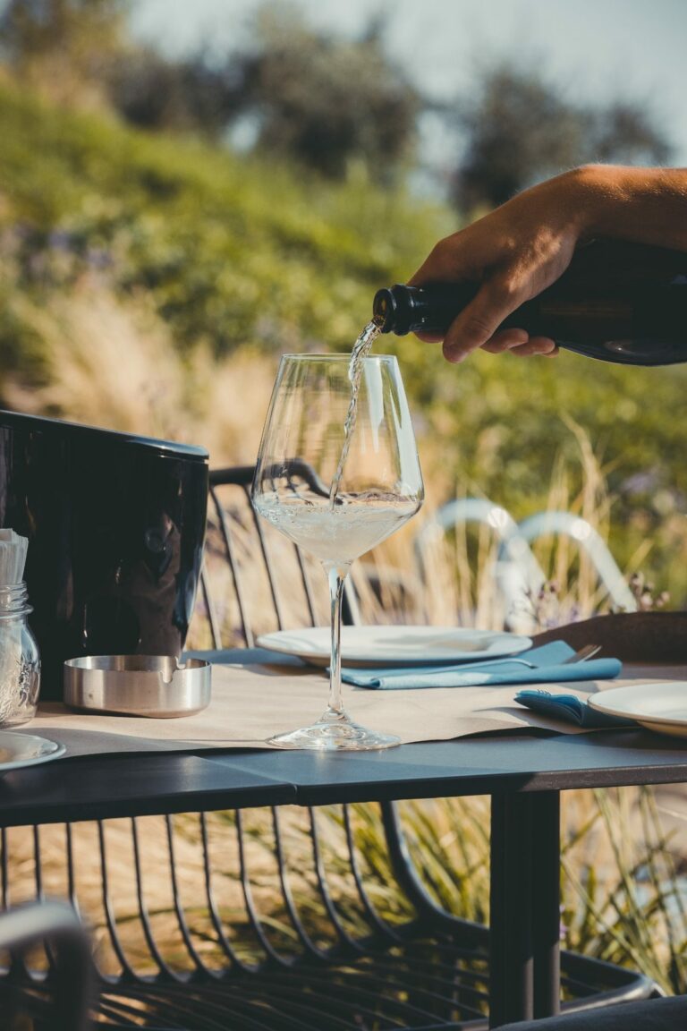 Une main verse du vin blanc dans un verre posé sur une table en terrasse dressée avec des assiettes, des couverts et un seau à vin, le tout sur fond de verdure floue où se détache l'élégance d'un camion-bar événementiel.