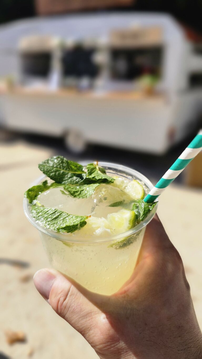 Une main tient un verre transparent rempli de limonade, avec des glaçons, des feuilles de menthe et une paille rayée verte. À l'arrière-plan, un camion-bar événementiel flou contribue à l'ambiance animée.